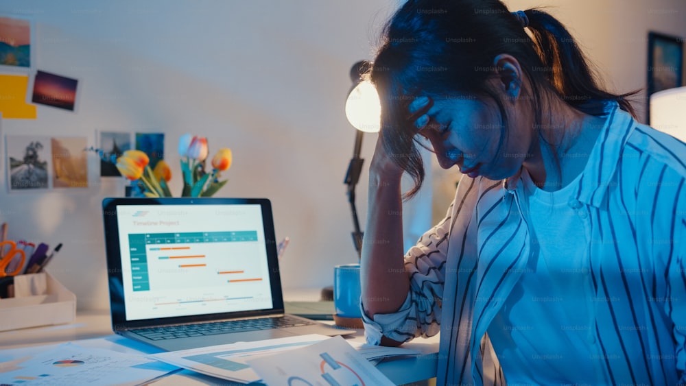 Joven empleada asiática trabaja horas extras a altas horas de la noche estrés finanzas finanzas investigación de proyectos en computadora portátil en casa. Las estudiantes aprenden en línea en casa, distancia social, concepto de trabajo normal desde casa.
