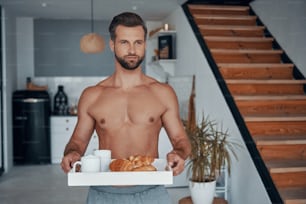 Good looking young shirtless man holding tray with breakfast while spending time at home
