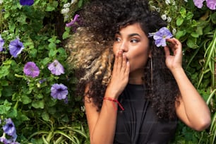 Attractive tender young woman with afro hairstyle posing over fresh natural flowers outdoor. Romantic surprised girl. Date. Real people emotions.