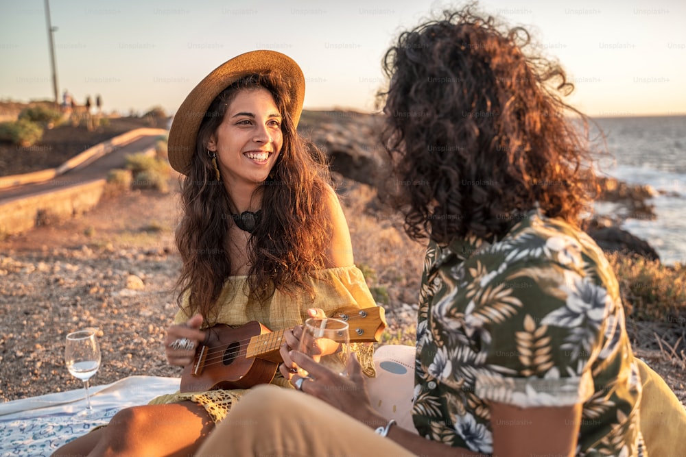 Young fashionable hippie couple on a romantic picnic on the beach, playing on ukulele guitar and singing songs. Sunset light. Summer time.