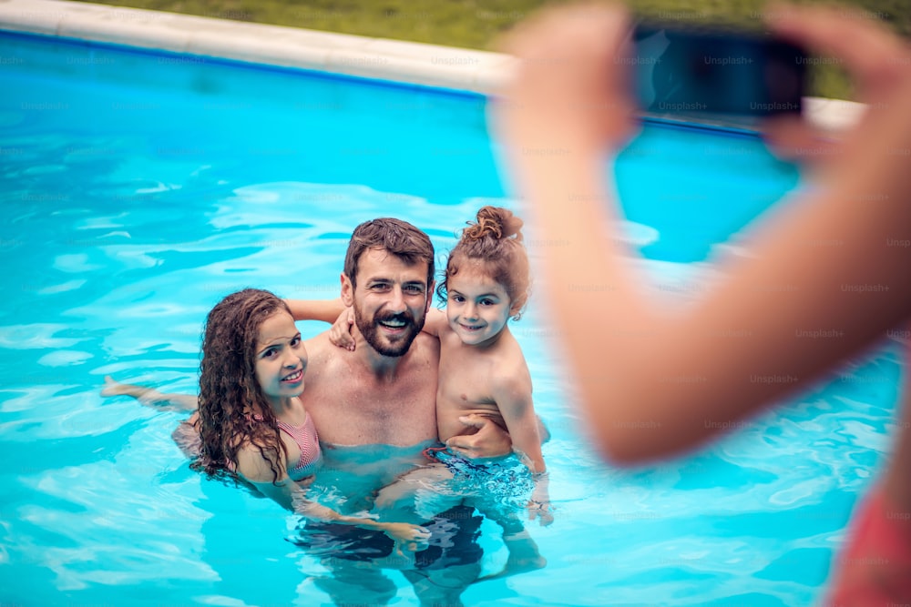 Father and children playing in the swimming pool. Woman taking photo of man and kids. Focus is on foreground.