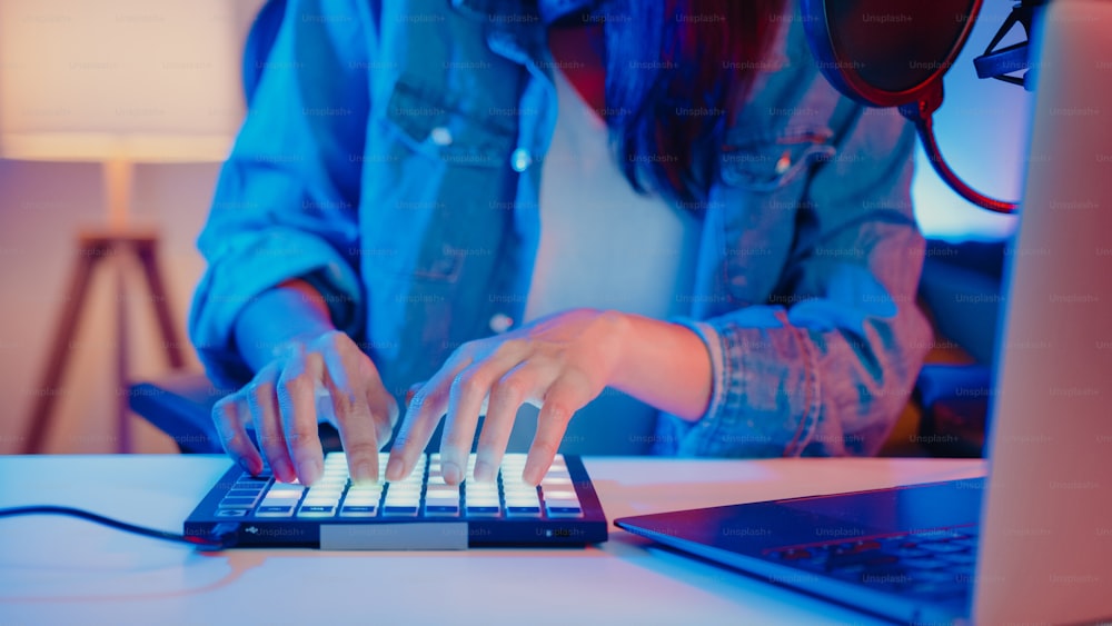 Primo piano felice ragazza asiatica dj uso launchpad sintetizzatore tastiera suono mixer indossare cuffia fatto musica in computer portatile online dal vivo in soggiorno home studio di notte. Concetto di creatore di contenuti.