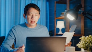 Junger asiatischer Geschäftsmann konzentriert Online-Videoanruf-Meeting-Auftrag auf Papierkram mit Kollegen in Laptop-Computer im Wohnzimmer zu Hause Überstunden in der Nacht, Arbeit von zu Hause aus Corona-Pandemie-Konzept.