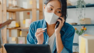 Junge asiatische Geschäftsfrauen tragen Gesichtsmasken mit Handyanruf, um Bestellungen zu erhalten und Produkte auf Lagerarbeit im Heimbüro zu überprüfen. Kleinunternehmer, Online-Marktlieferung freiberufliches Konzept.