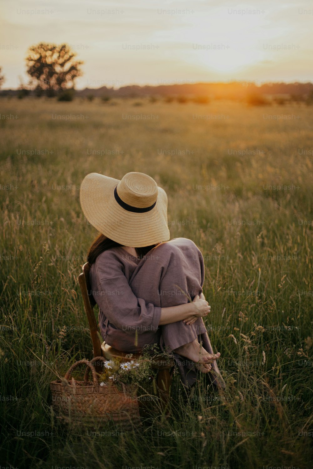 Schöne Frau in Leinenkleid und Strohhut sitzt auf rustikalem Stuhl auf Sommerwiese bei Sonnenuntergang. Junge Frau mit Blumenkorb entspannt sich auf dem Land. Atmosphärisch ruhiger, ruhiger Moment