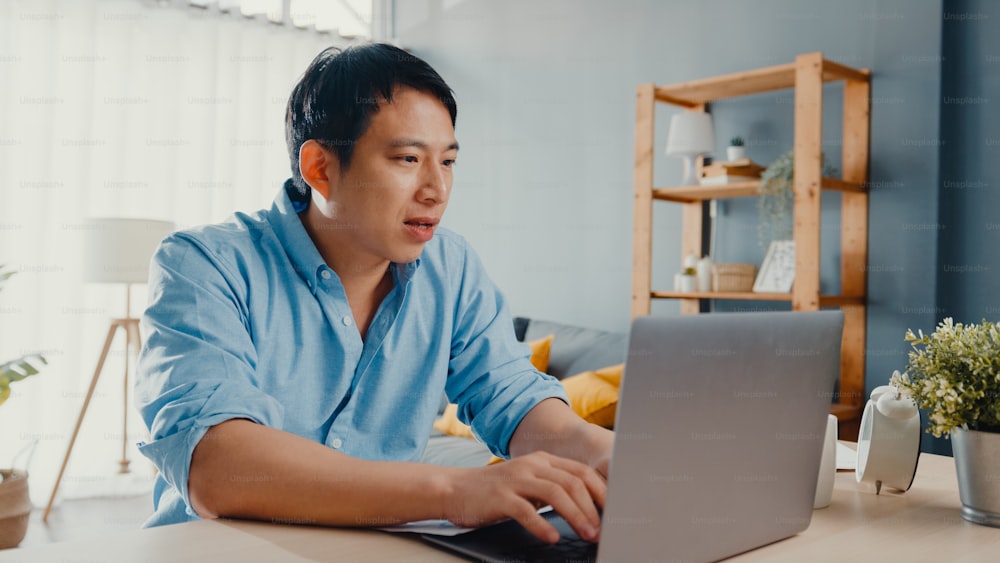 Freelance Asia guy casual wear using laptop online learning in living room at house. Working from home, remotely work, distance education, social distancing, quarantine for corona virus prevention.