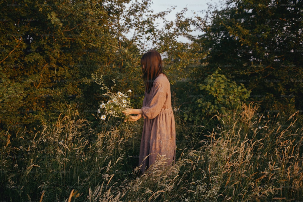 Mujer hermosa en vestido de lino recogiendo flores silvestres en el prado de verano en la tarde soleada. Mujer joven con estilo en vestido rústico recogiendo flores en el campo. Momento rural atmosférico