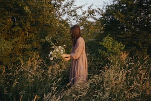 Mujer hermosa en vestido de lino recogiendo flores silvestres en el prado de verano en la tarde soleada. Mujer joven con estilo en vestido rústico recogiendo flores en el campo. Momento rural atmosférico