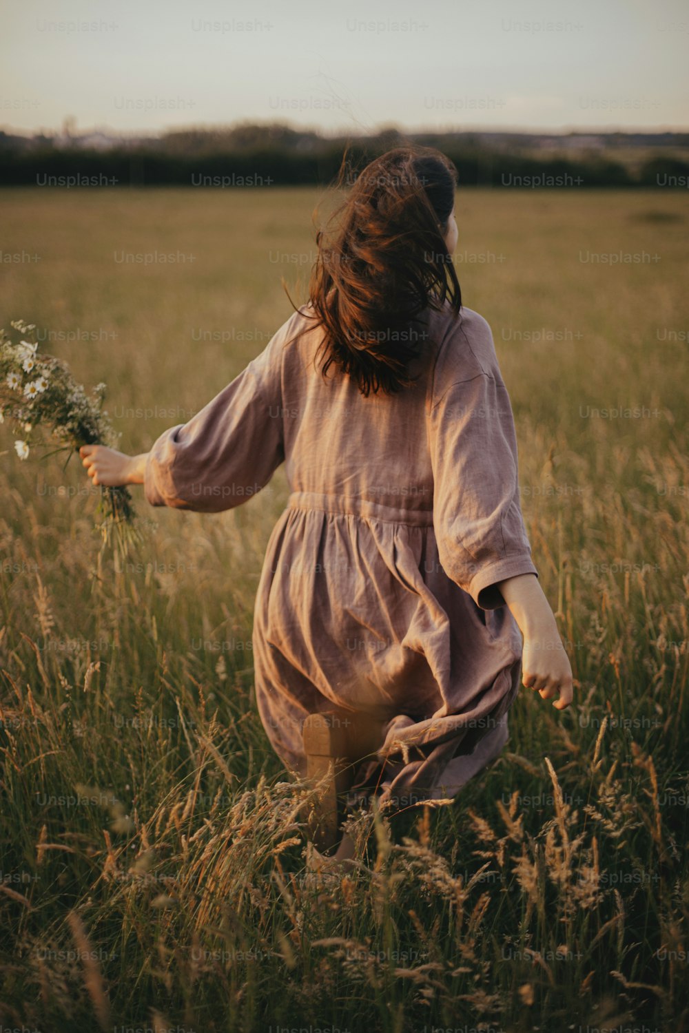 Beautiful woman in linen dress running with wildflowers in hand in summer meadow in sunset. Stylish young female in rustic dress enjoying free evening in countryside. Atmospheric carefree moment