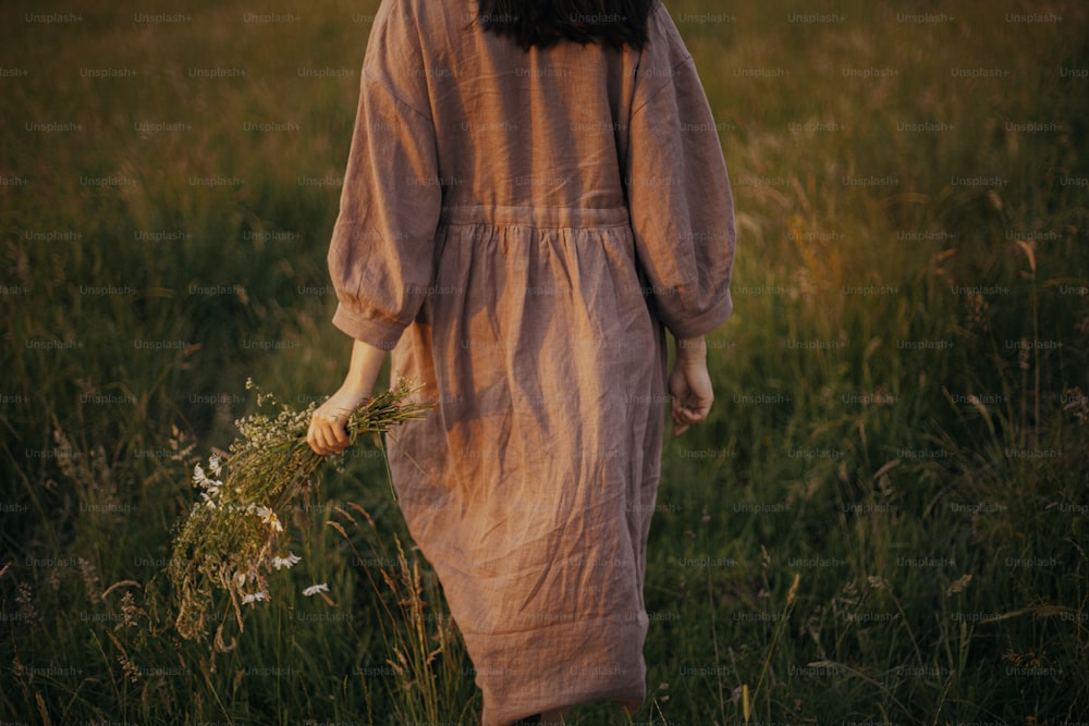 Beautiful woman in linen dress walking with wildflowers in hand in summer meadow in sunset, back view. Young female in rustic dress relaxing in evening in countryside. Atmospheric calm moment