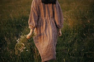 Beautiful woman in linen dress walking with wildflowers in hand in summer meadow in sunset, back view. Young female in rustic dress relaxing in evening in countryside. Atmospheric calm moment