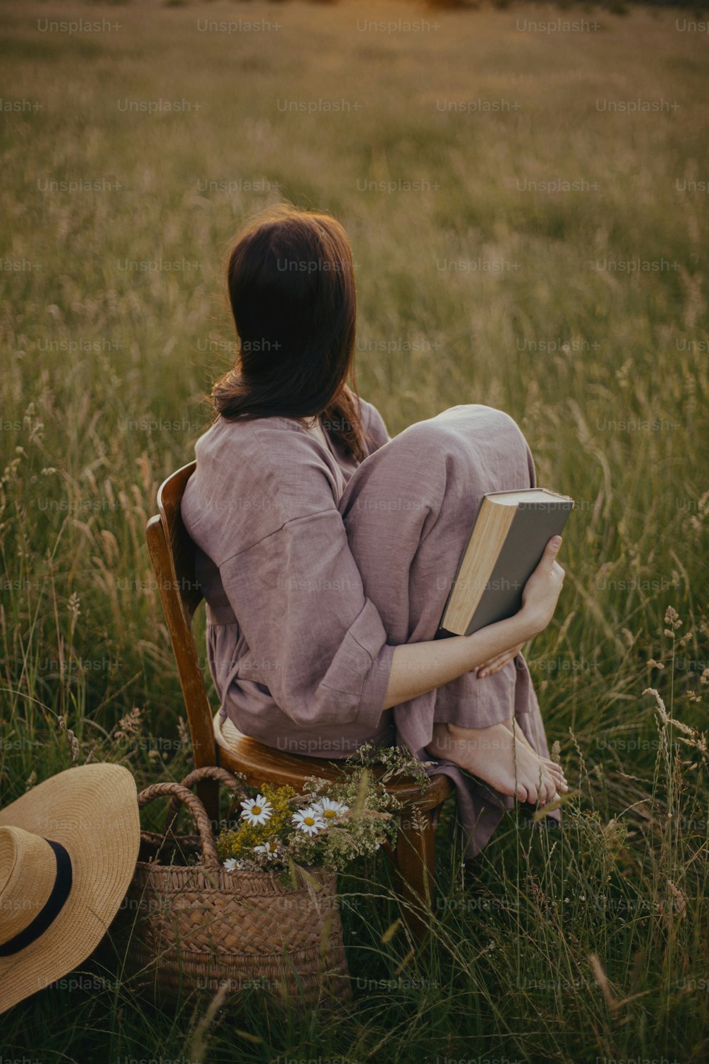 Schöne Frau im Leinenkleid, die auf rustikalem Stuhl sitzt und den Sonnenuntergang auf der Sommerwiese genießt. Junge Frau entspannt sich mit Buch und Blumenkorb auf dem Land. Atmosphärischer ruhiger Moment