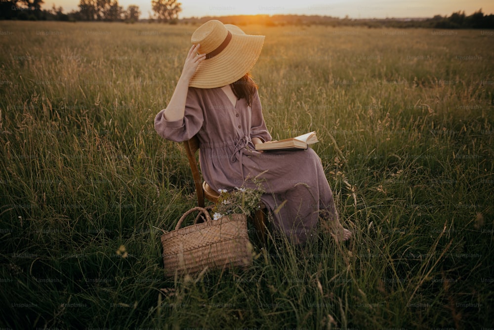 Schöne Frau in Hut und Leinenkleid mit Buch und Blumenkorb auf rustikalem Stuhl auf Sommerwiese bei Sonnenuntergang. Atmosphärischer ruhiger Moment. Junge Frau liest Buch auf dem Land