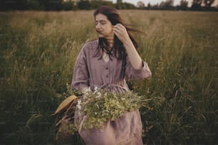 Beautiful woman with windy hair and in linen dress sitting on wooden chair with bouquet of flowers in summer meadow. Young female relaxing in evening countryside. Atmospheric carefree moment