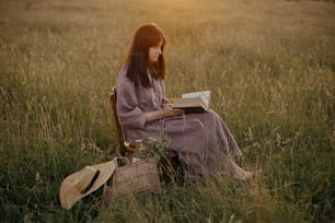 Bella donna in abito di lino con libro e cesto di fiori seduta su sedia di legno nel prato estivo al tramonto. Momento di calma atmosferica. Giovane femmina che si rilassa nella campagna serale