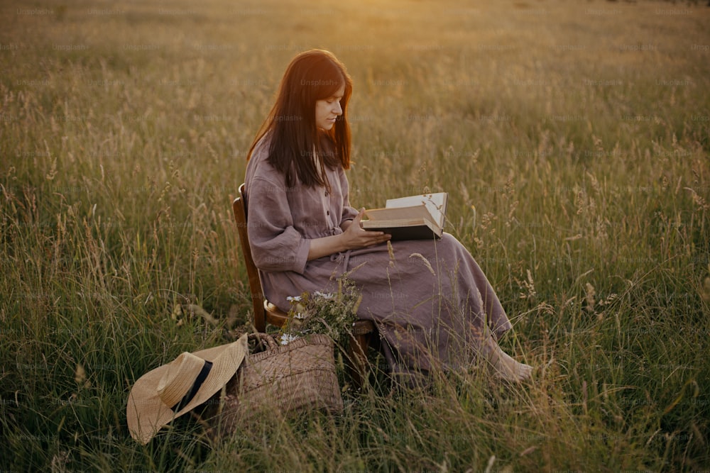 Beautiful woman in linen dress with book and basket of flowers sitting on wooden chair in summer meadow in sunset. Atmospheric calm moment. Young female relaxing in evening countryside