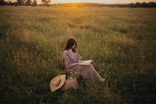 Mulher bonita no vestido de linho com livro e cesta de flores sentadas na cadeira de madeira no prado de verão no pôr do sol. Fêmea nova relaxando no campo da noite. Momento de calma atmosférica
