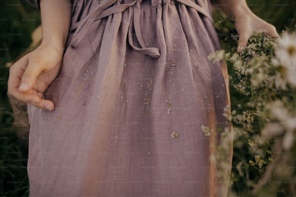 Beautiful linen dress with petals of wildflowers on woman among grass in summer meadow, cloth close up. Gathering daisy and cow parsley in countryside. Atmospheric moment