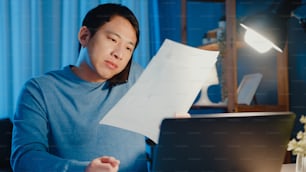 Young Asia businessman use smartphone call meeting agenda assignment paperwork with colleague look at laptop computer in living room at home overtime at night, Work from home corona pandemic concept.