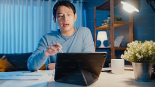 Asia businessman focus online video call meeting assignment on paperwork with colleague in tablet computer in living room at home overtime at night, Work from home coronavirus pandemic concept.