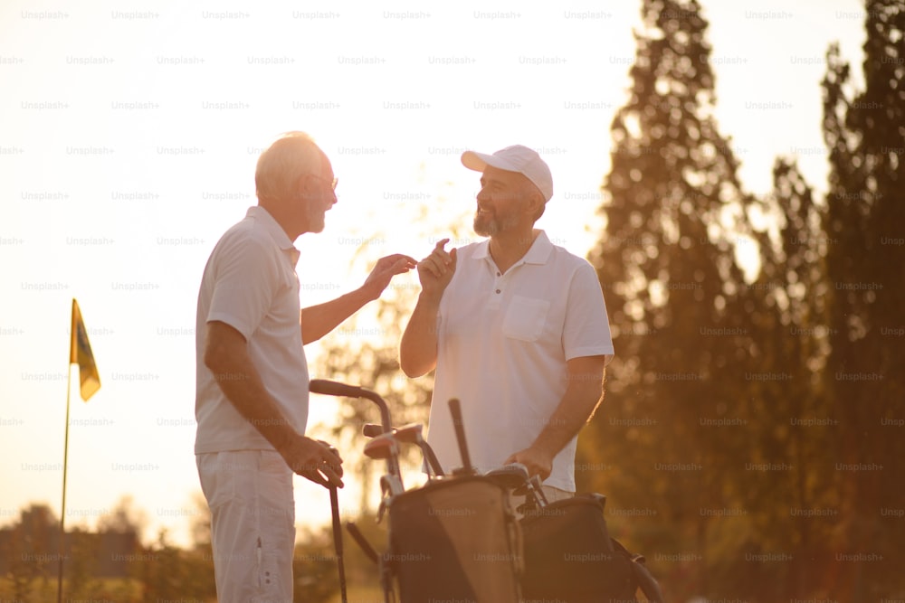 Two older men stand on a golf course and talk.