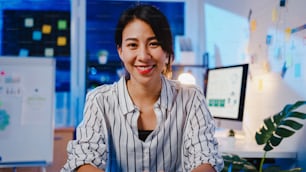 Portrait of beautiful executive businesswoman smart casual wear looking at camera and smiling, happy in modern office workplace night. Young Asia lady talk to colleague in video call meeting at home.