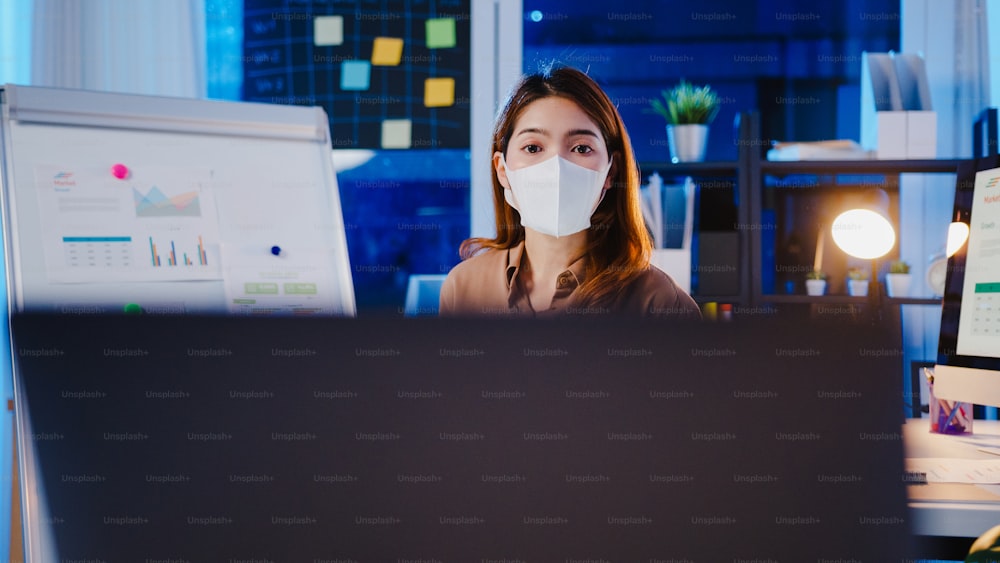 Empresária asiática usa máscara facial para distanciamento social no novo normal para apresentação de prevenção ao vírus ao colega sobre plano em chamada de vídeo enquanto trabalha no escritório à noite. Estilo de vida após o coronavírus.