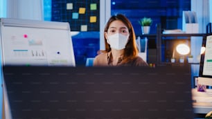 아시아 사업가는 사무실 밤에 일하는 동안 화상 통화로 계획에 대해 동료에게 바이러스 예방 프레젠테이션을 위해 뉴 노멀에서 사회적 거리두기를 위해 안면 마스크를 착용합니다. 코로나 바이러스 이후의 라이프 스타일.