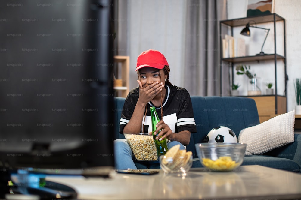Mujer afroamericana emocionada con atuendo informal bebiendo cerveza y comiendo bocadillos mientras ve un partido de fútbol en la televisión. Concepto de personas, deporte y tiempo libre.