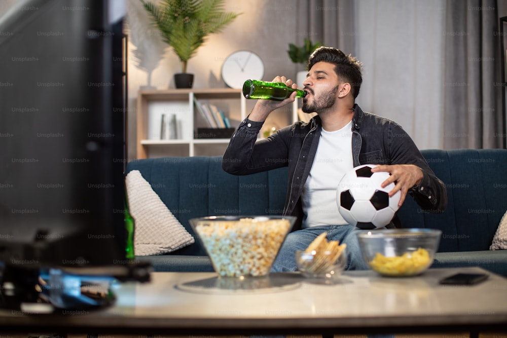 Cara indiano elegante bebendo cerveja gelada enquanto se senta confortavelmente no sofá e desfruta do campeonato de futebol em casa. Conceito de tempo livre, esporte e entretenimento.