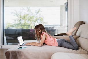 Menina bonita deita-se no sofá da sala de estar e usa um laptop para surfar, aprender e comunicação. Ela está feliz e sorriu.