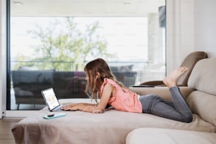 Beautiful girl lies on the living room sofa and uses a laptop for surfing, learning and communication. She is happy and smiled.