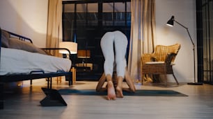 Young Asia lady in sportswear exercises doing work out and using tablet to watch yoga video tutorial at home night. Distant training with personal trainer, social distance, online education concept.