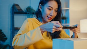 Una mujer asiática joven usa un teléfono inteligente para tomar una imagen del código de barras en el producto del paquete para la entrega del envío al cliente en la oficina en casa por la noche. Pequeña empresa, entrega de mercado en línea, concepto independiente de estilo de vida.