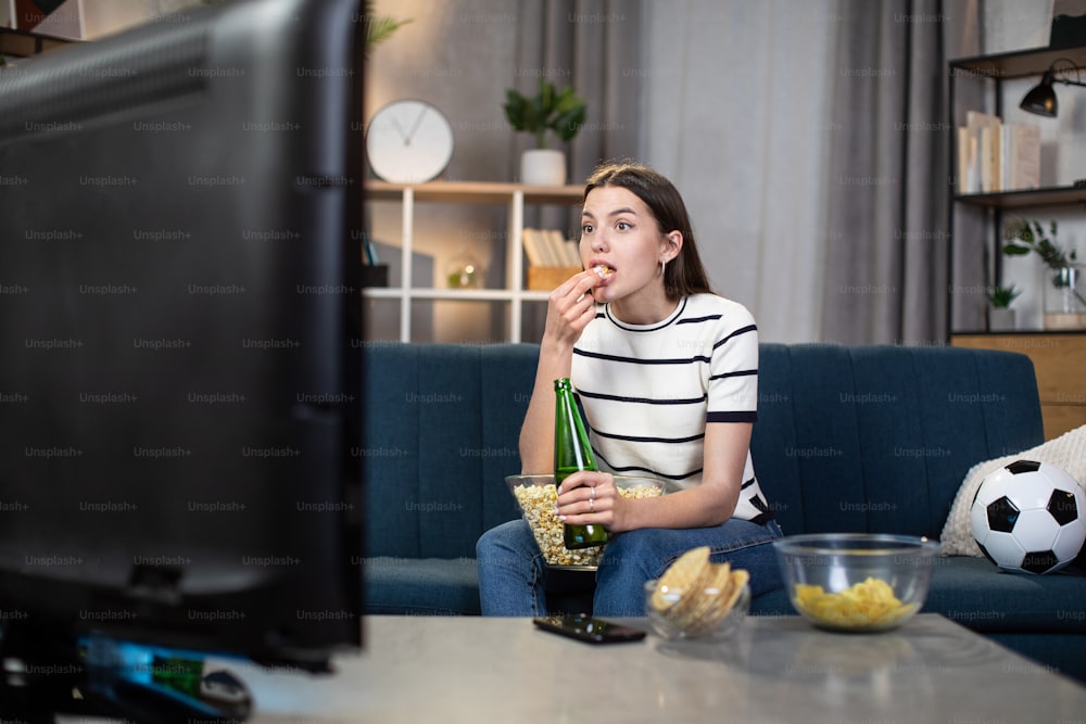 Bella giovane donna in abbigliamento casual che mangia snack e beve birra mentre guarda la partita di calcio in TV a casa. Concetto di tifoso, tempo libero e sport.