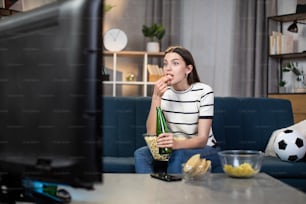 Beautiful young woman in casual wear eating snacks and drinking beer while watching football match on TV at home. Concept of fan, leisure and sport.