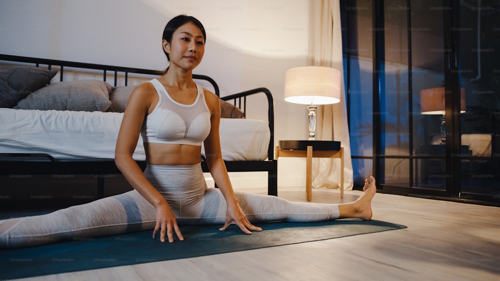 Jeune femme asiatique en vêtements de sport faisant de l’exercice de yoga dans le salon à la maison la nuit. Activité sportive et récréative, distanciation sociale, quarantaine pour le concept de prévention du virus corona.