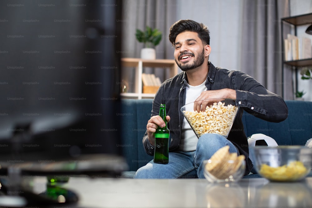 Young bearded man eating snacks and drinking cold beer while watching football game at home. Indian soccer fan sitting on couch and enjoying sport competition.
