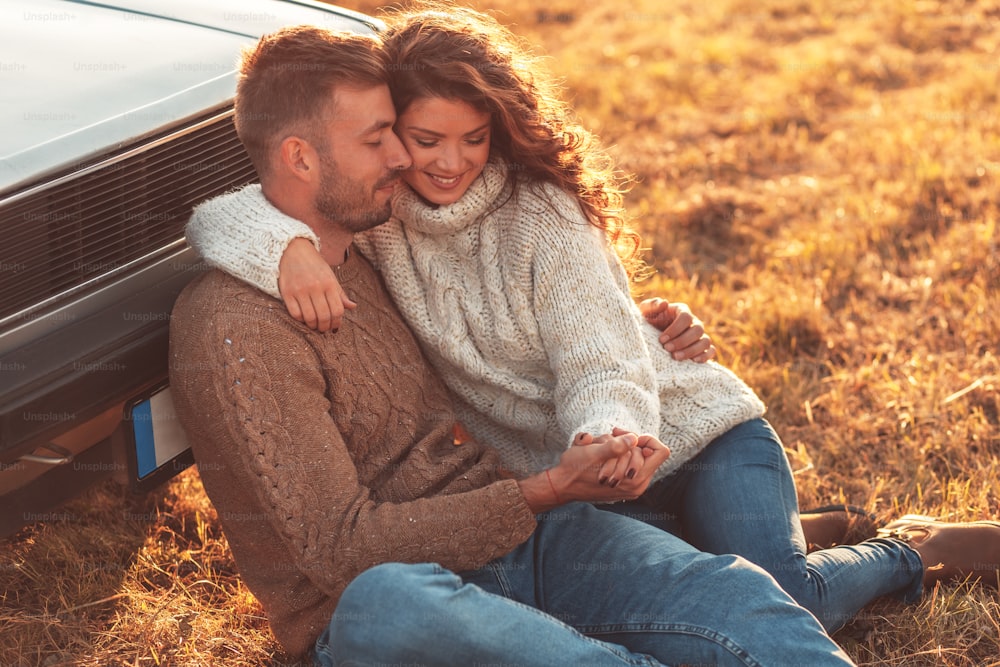 美しい若いカップルは、お互いに抱き合って昔ながらの車にもたれかかって牧草地に座って、屋外で一緒に時間を楽しんでいます。