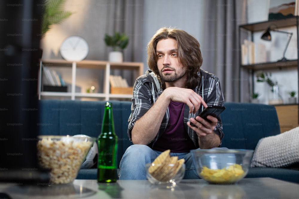 Junger kaukasischer Mann in Freizeitkleidung schaut Sportprogramm im Fernsehen, während er auf der Couch sitzt und ein modernes Smartphone in den Händen hält. Menschen und Technik konzeptieren.