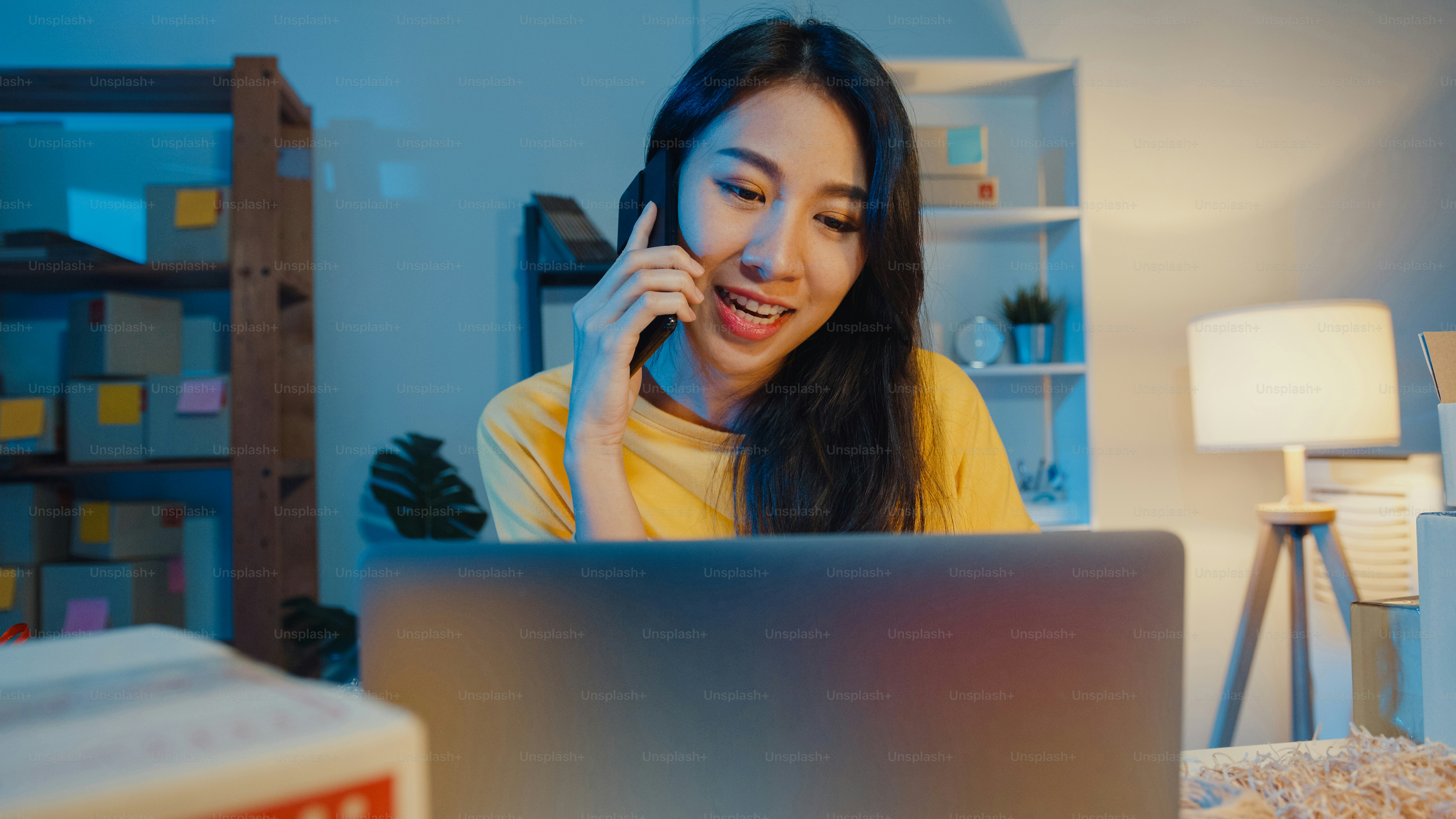 Young Asia woman call smartphone talk with customer for check confirm order in stock on laptop computer in home office at night. Small business, online market delivery, lifestyle freelance concept.