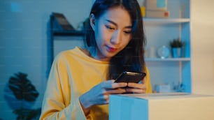 Una mujer asiática joven usa un teléfono inteligente para tomar una imagen del código de barras en el producto del paquete para la entrega del envío al cliente en la oficina en casa por la noche. Pequeña empresa, entrega de mercado en línea, concepto independiente de estilo de vida.