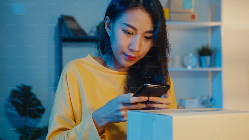 Young Asia woman use smartphone take barcode picture on parcel product for shipping delivery to customer in home office at night. Small business , online market delivery, lifestyle freelance concept.