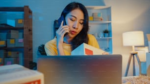 Junge Asia-Frau rufen Smartphone-Gespräch mit Kunden für Scheck bestätigen Bestellung auf Lager auf Laptop-Computer im Heimbüro in der Nacht. Kleinunternehmen, Online-Marktlieferung, Lifestyle-Freelance-Konzept.