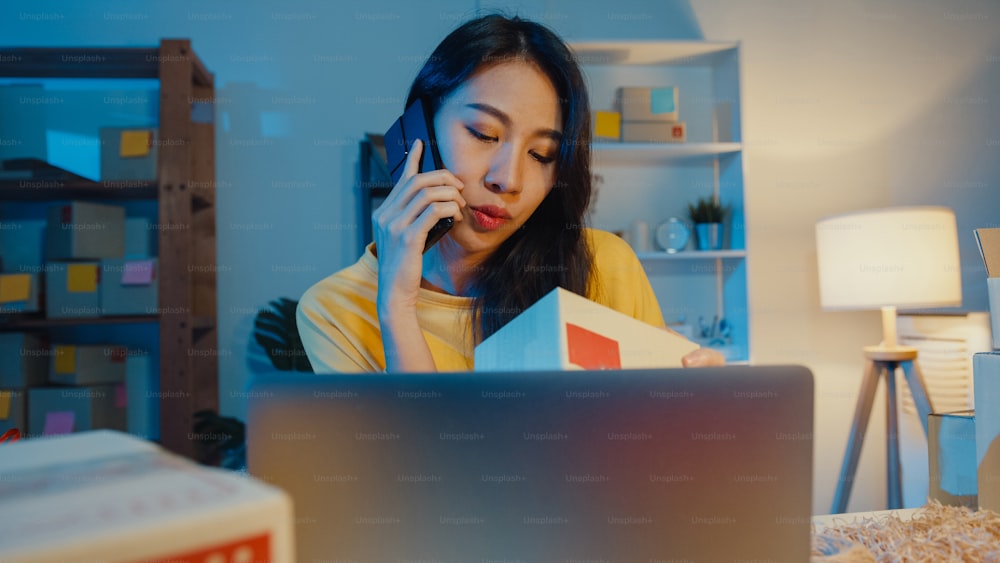 Young Asia woman call smartphone talk with customer for check confirm order in stock on laptop computer in home office at night. Small business, online market delivery, lifestyle freelance concept.