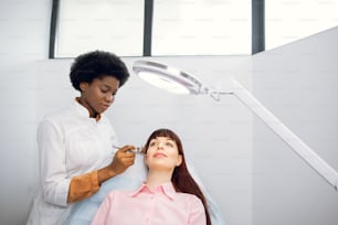 Bio revitalization and oxygen peeling therapy. Young lady in the beauty salon during rejuvenation procedure. African female beautician using the apparatus, sprayes a cosmetic product on patient's skin
