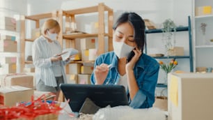 Young Asia businesswomen wear face mask using mobile phone call receiving purchase order and check product on stock work at home office. Small business owner, online market delivery freelance concept.