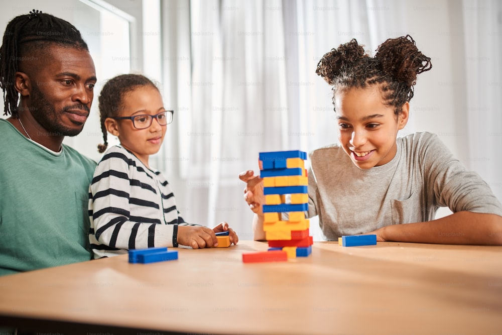 Waist up portrait view of the multiracial girl pulls a bar while playing at the jenga with her father and sister at the kitchen. Stock photo