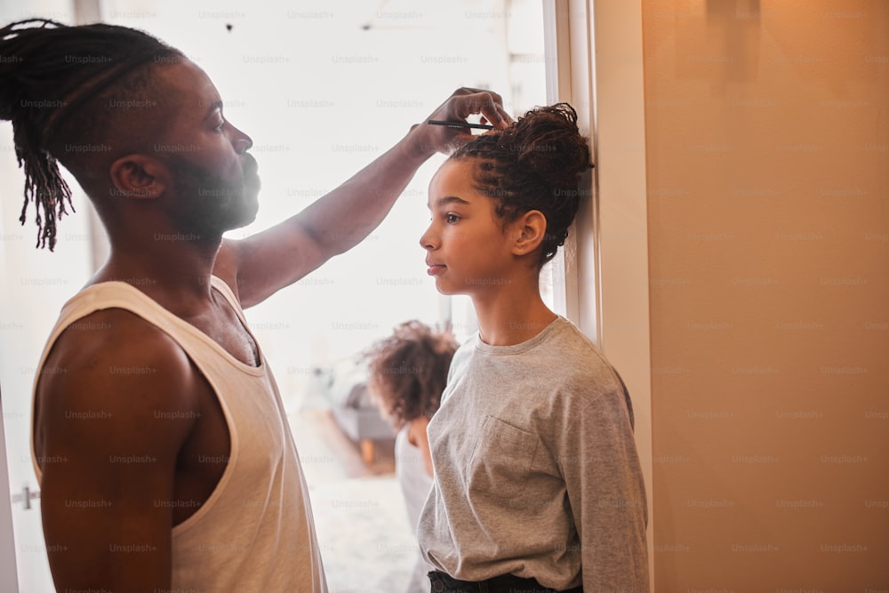 What is my height. Waist up portrait view of the strong man with dreadlocks measuring height of his teenage daughter at home. Stock photo