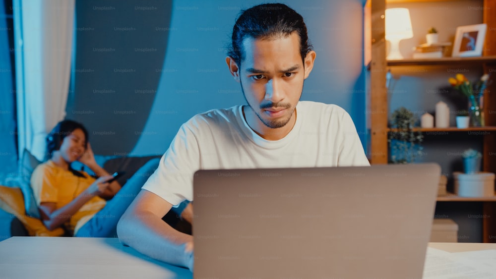 Freelance asian couple man and woman in casual husband focus work in laptop computer at night wife take a rest feel relax wear headphones play music on smartphone lay down and dance on couch at home.